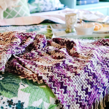 A woven purple throw blanket draped across a bed. 