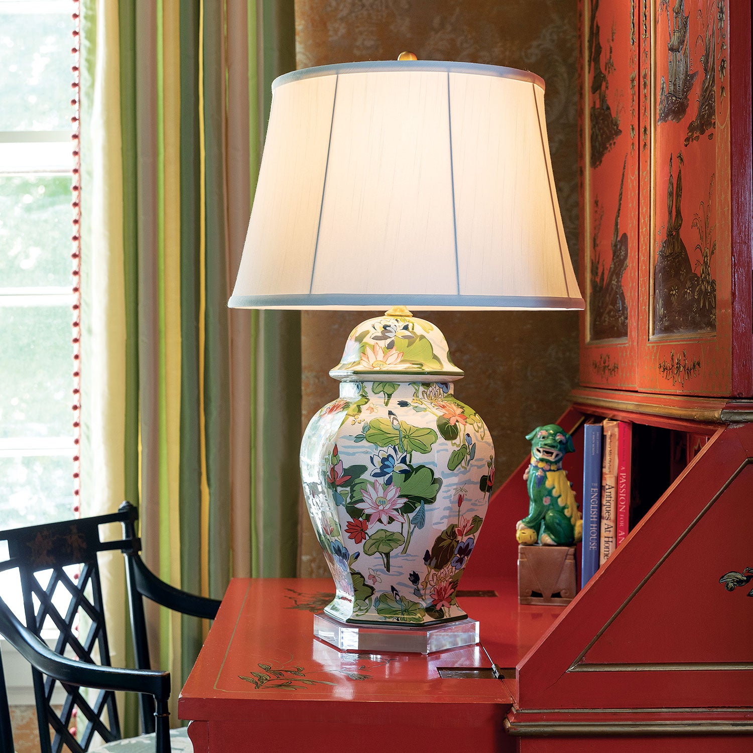 A porcelain buffet lamp covered in waterlily motifs with a plain white lampshade sitting on a red bureau.