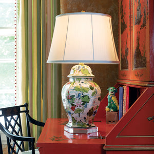 A porcelain buffet lamp covered in waterlily motifs with a plain white lampshade sitting on a red bureau. 