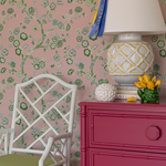 chest, lamp and chair against pink and green chinoiserie wallpaper featuring green cherry blossom motifs on a pink  background. 