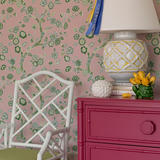 chest, lamp and chair against pink and green chinoiserie wallpaper featuring green cherry blossom motifs on a pink  background. 