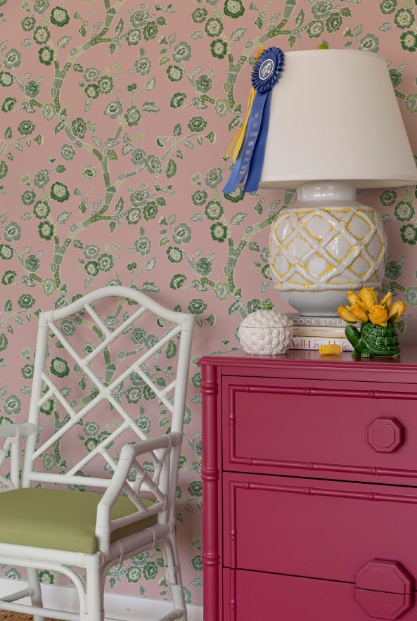 chest, lamp and chair against pink and green chinoiserie wallpaper featuring green cherry blossom motifs on a pink  background. 