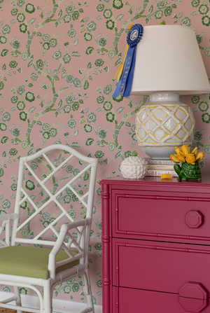 chest, lamp and chair against pink and green chinoiserie wallpaper featuring green cherry blossom motifs on a pink  background. 