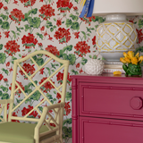 Red geranium wallpaper in a room featuring a pink chest of drawers with a large lamp and a yellow bamboo chair.