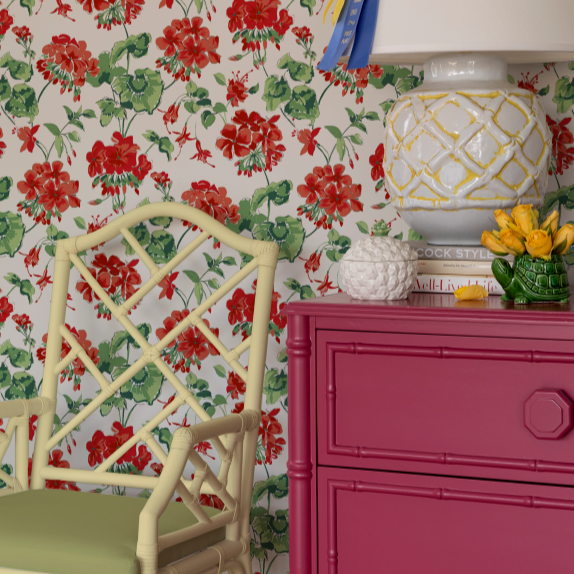 Red geranium wallpaper in a room featuring a pink chest of drawers with a large lamp and a yellow bamboo chair. 