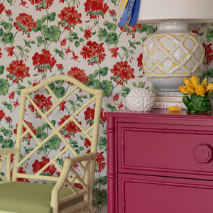 Red geranium wallpaper in a room featuring a pink chest of drawers with a large lamp and a yellow bamboo chair. 