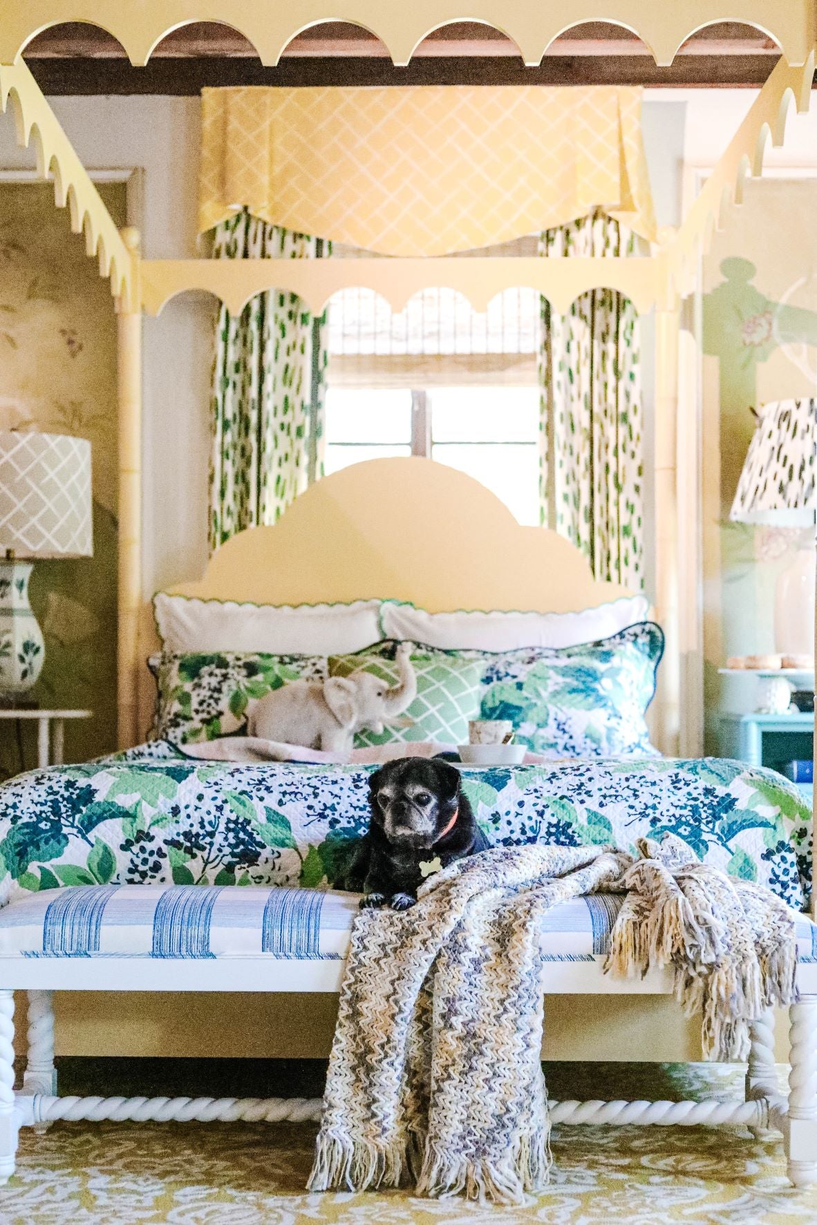 a yellow four poster canopy bed with floral bedding and a black pug dog on a bench at the end. 